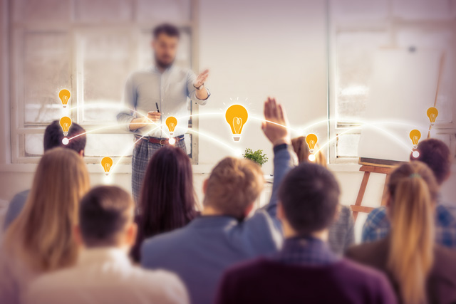 Ein Dozent steht vor einer Gruppe. Ein Mann meldet sich. Das Bild steht für eine wichtige Eigenschaft der digitalen Baugenehmigung. Sie wurde von der Verwaltung auch für die einfachere Verwaltung entwickelt.